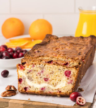 partially sliced cranberry cream cheese bread on a wooden cutting board with cranberries and oranges in the background