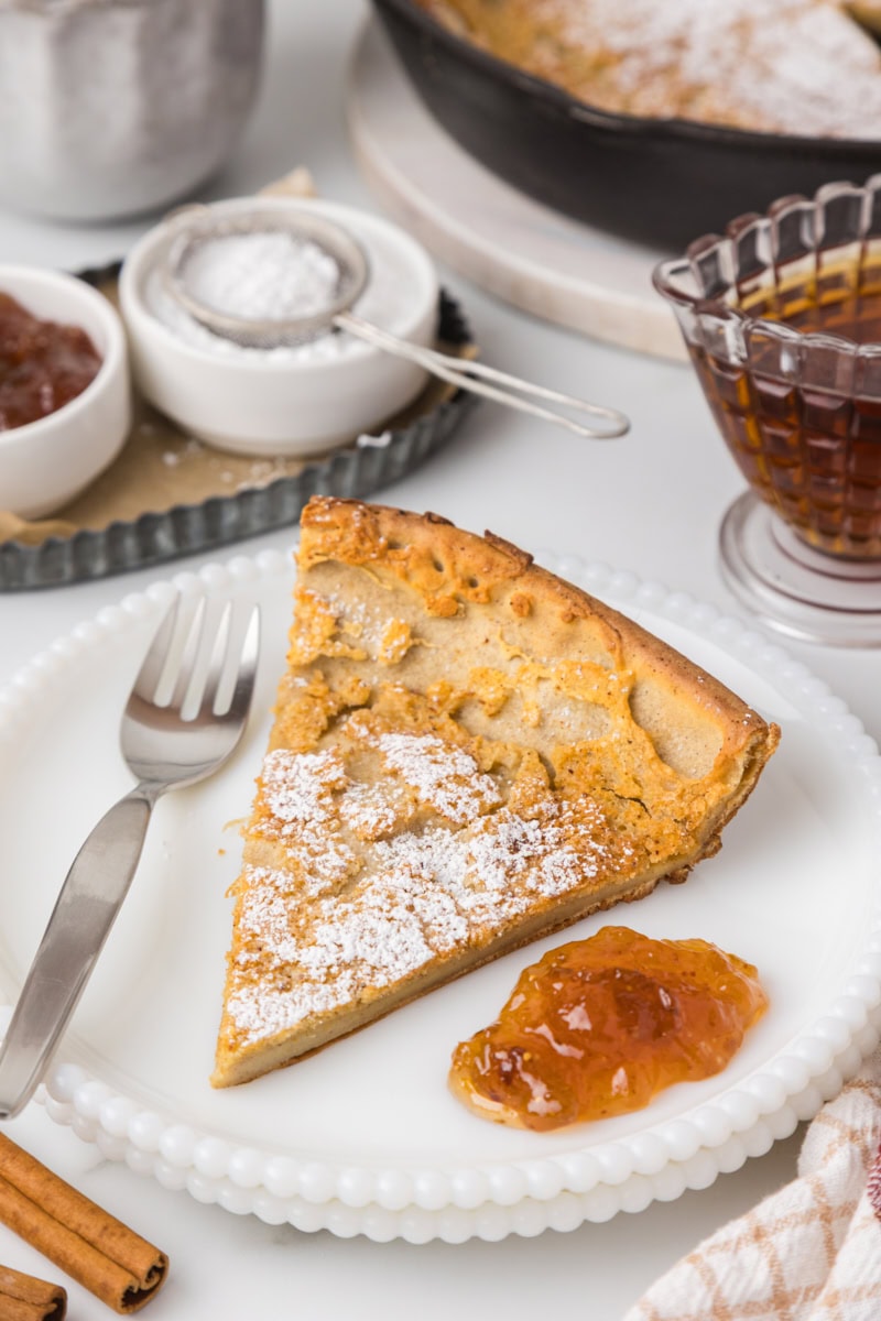 a slice of Dutch baby pancake dusted with powdered sugar and served with a dollop of jam on a white plate