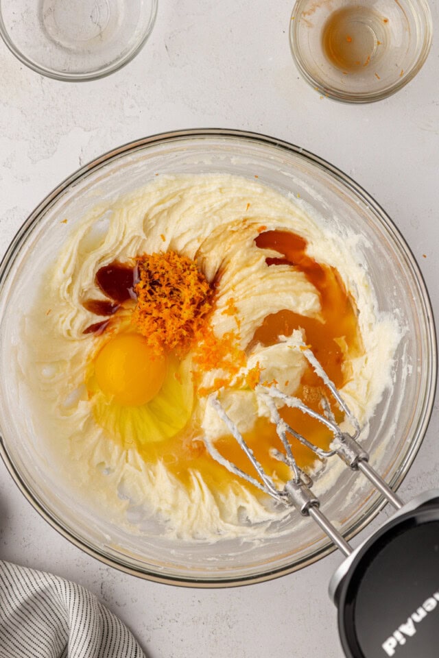 overhead view of egg, orange zest, orange juice, vanilla, and almond extract added to creamed butter, cream cheese, and sugar in a mixing bowl
