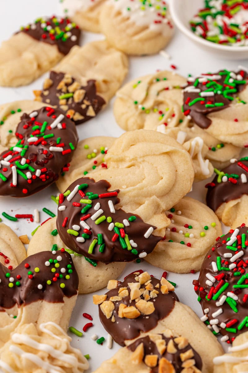 whipped shortbread cookies scattered on a white surface, showing a variety of shapes and decor including s-shapes, rounds, sprinkles, and chocolate-dipped