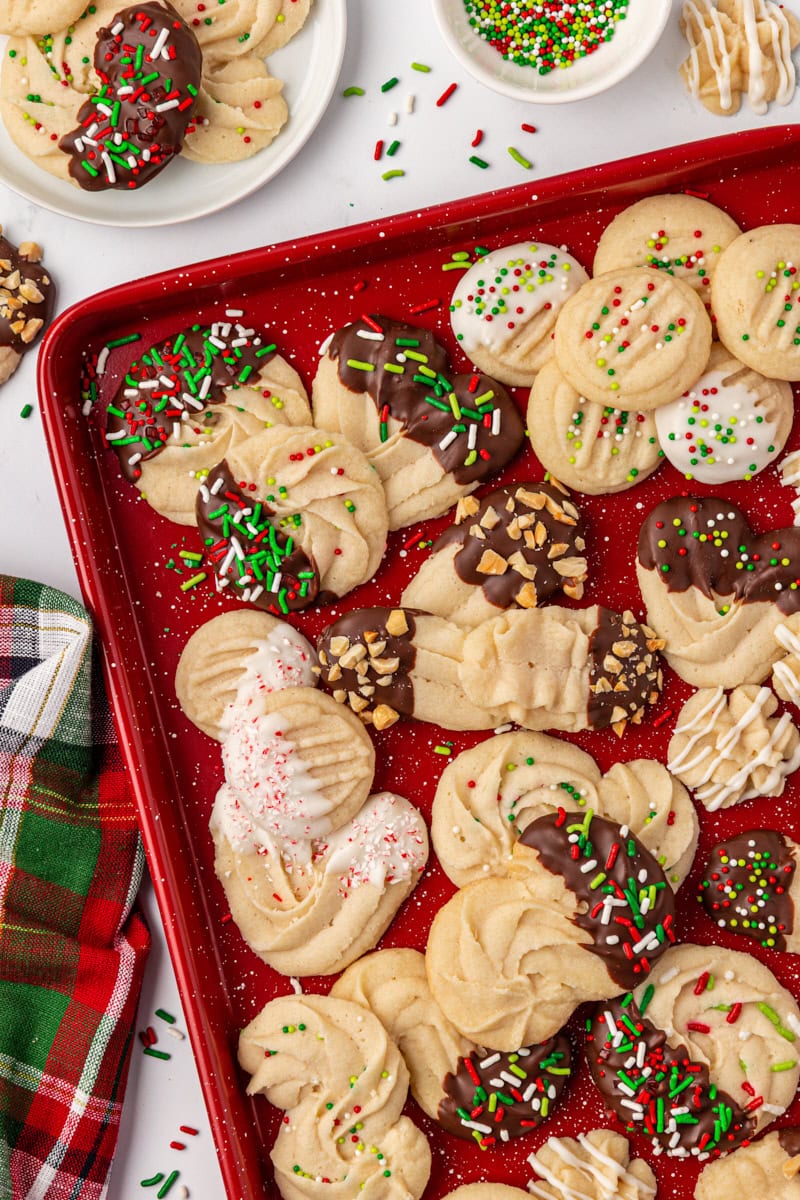 Whipped shortbread cookies arranged on a red baking sheet, showing a variety of shapes and decorations like wreaths, S-shapes, chocolate dips, and sprinkles.