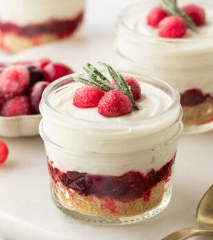 a no-bake white chocolate cranberry cheesecake jar on a white surface with more cheesecake jars and sugared cranberries in the background
