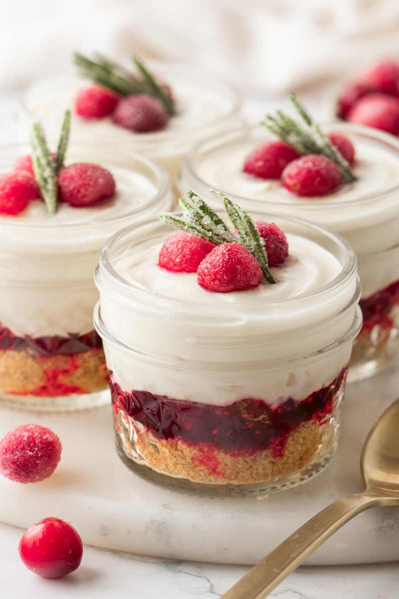 four no-bake white chocolate cranberry cheesecake jars topped with sugared cranberries and rosemary on a marble serving tray