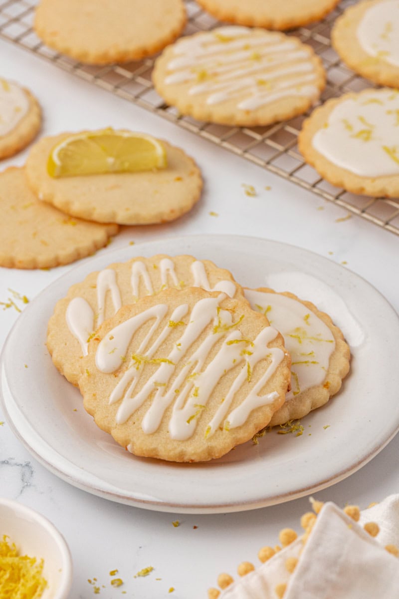 three lemon shortbread cookies on a white plate with more cookies in the background