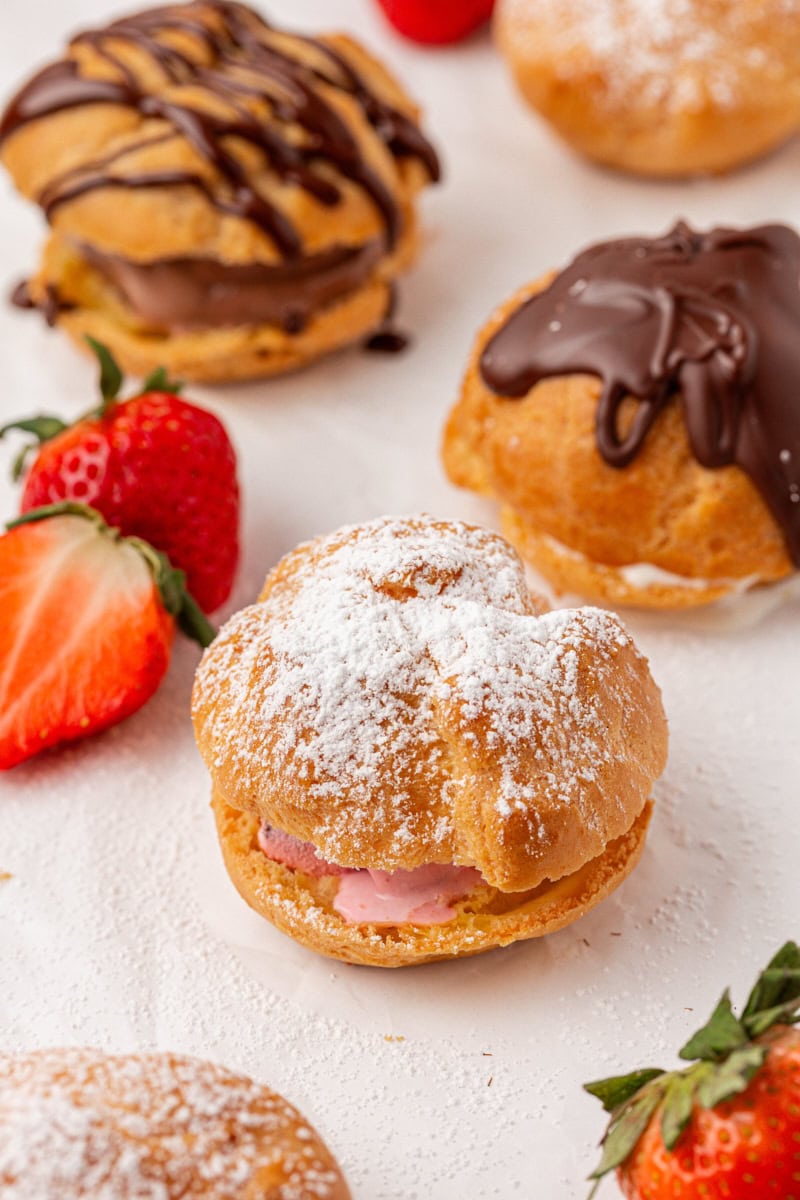 a profiterole filled with strawberry ice cream and topped with powdered sugar, with more profiteroles with various fillings and toppings surrounding