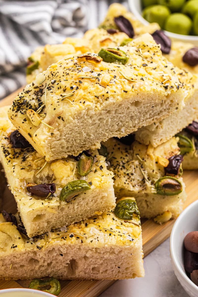 stacked pile of slices of no-knead focaccia topped with herbs, cheese, and olives