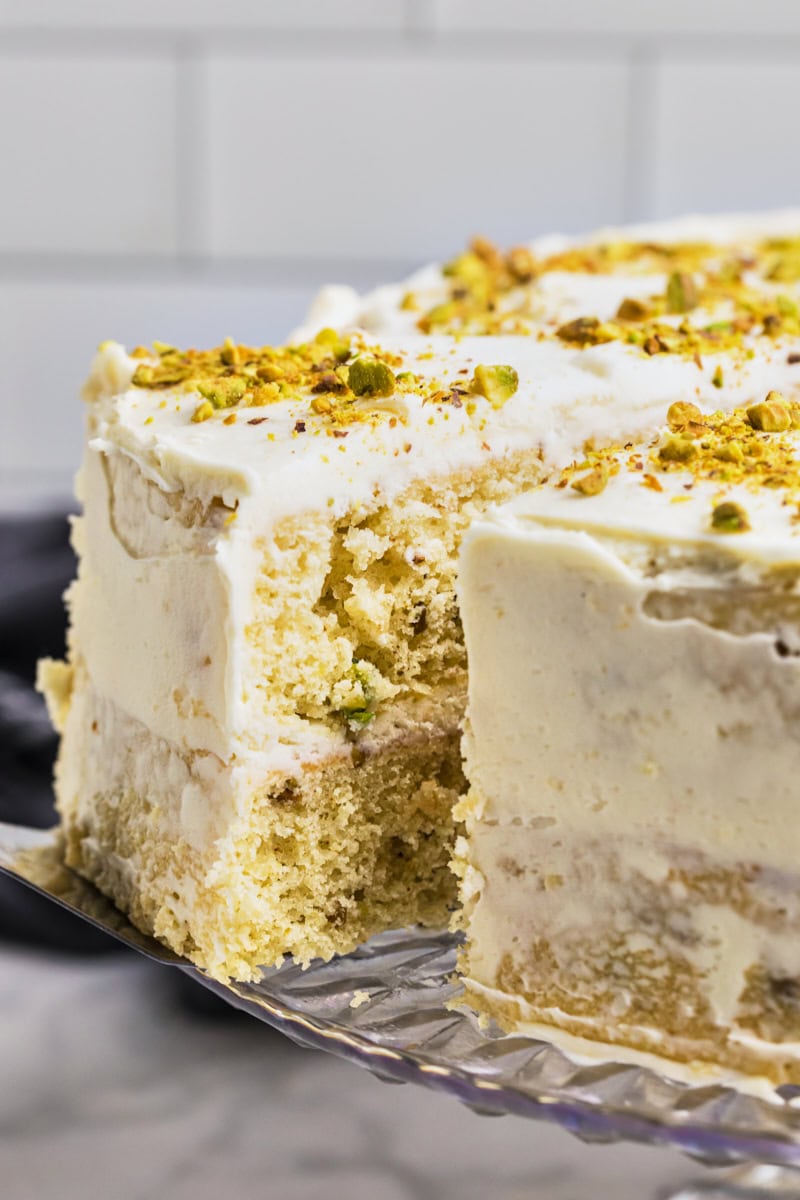 close up of a slice of pistachio cake with white chocolate frosting on a cake server being pulled away from the remaining cake