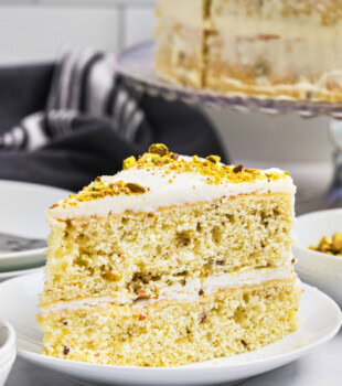 a slice of pistachio layer cake topped with white chocolate frosting with the remaining cake in the background
