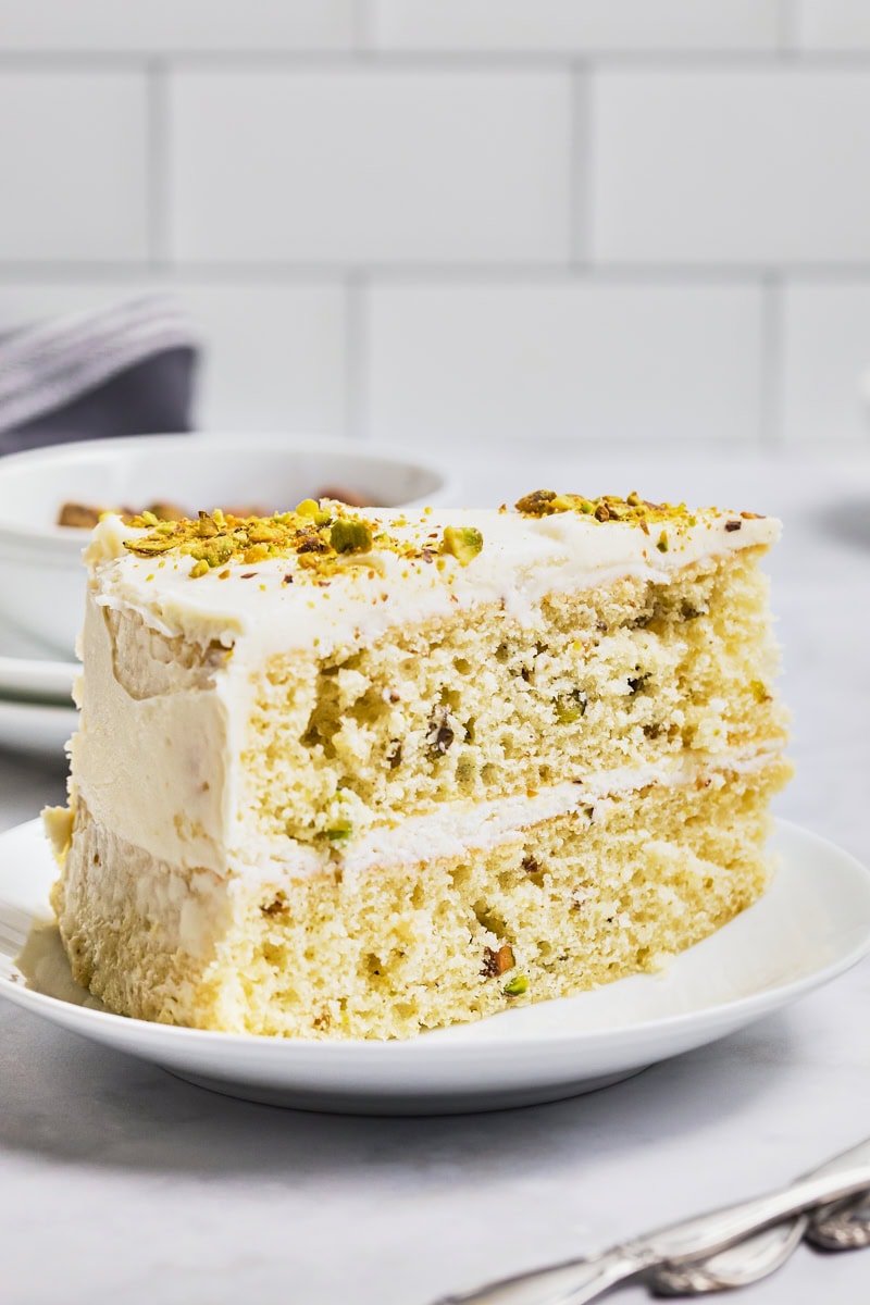 a slice of pistachio cake with white chocolate frosting and topped with chopped pistachios