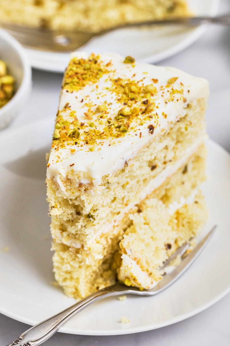 a slice of pistachio cake with white chocolate frosting on a white plate with a bite on the fork