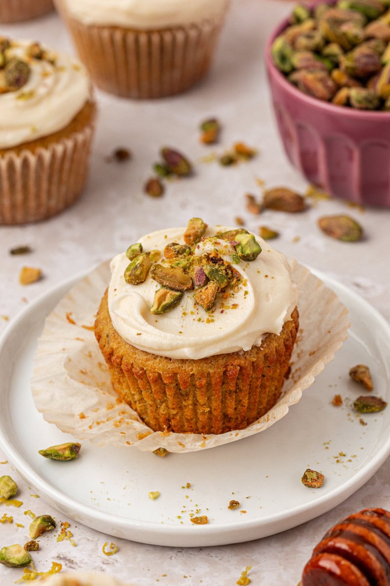 a partially unwrapped pistachio cupcake with honey cream cheese frosting on a white plate