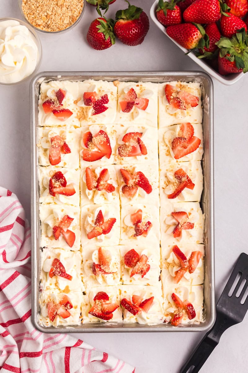 overhead view of strawberry shortcake cheesecake bars cut into squares and topped with whipped cream, fresh strawberries, and cookie crumbs