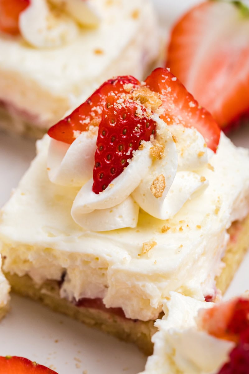 close up of a strawberry shortcake cheesecake bar topped with whipped cream, fresh strawberries, and vanilla cookie crumbs