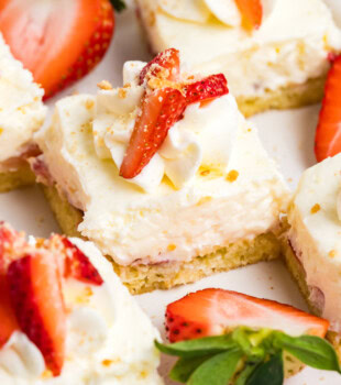 strawberry shortcake cheesecake bars featuring layers of vanilla cake, strawberries, and no-bake cheesecake filling topped with whipped cream, fresh strawberries, and vanilla cookie crumbs