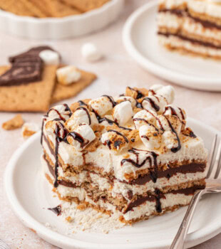 s'mores icebox cake with layers of marshmallow filling, graham crackers, and chocolate ganache, topped with chocolate sauce, cookie crumbs, and toasted marshmallows