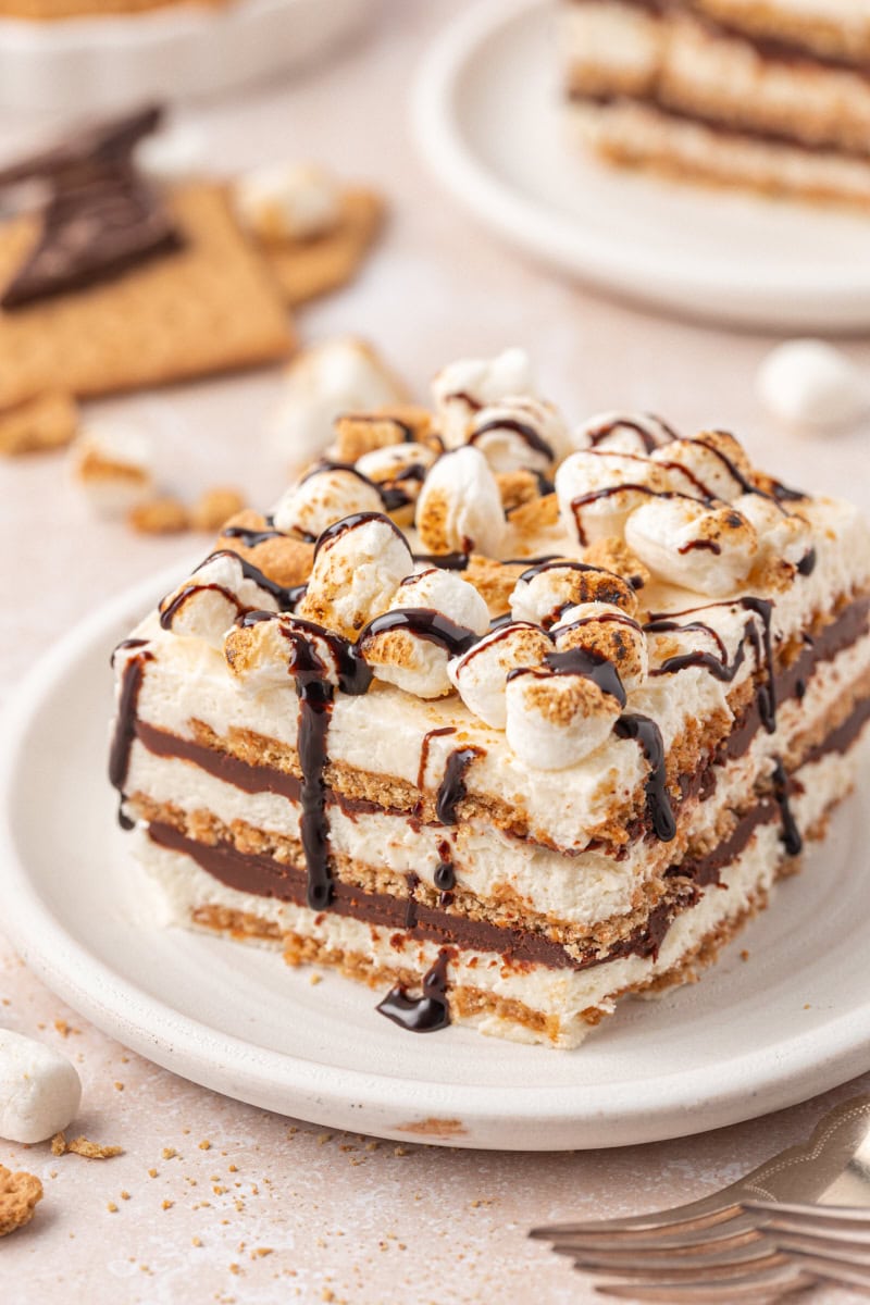 a slice of s'mores icebox cake with layers of graham crackers, marshmallow filling, and chocolate ganache, topped with toasted marshmallows, cookie crumbs, and chocolate sauce