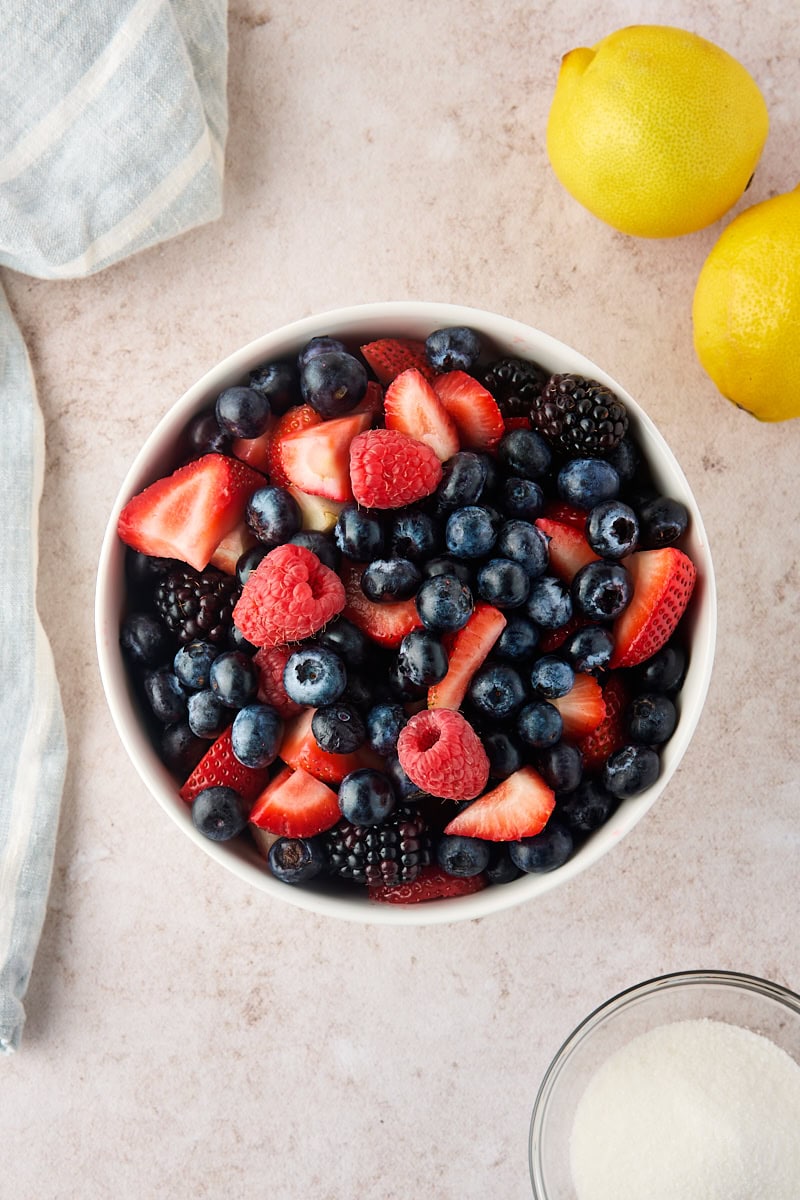 fresh mixed berries in a bowl, with lemons and a bowl of sugar alongside