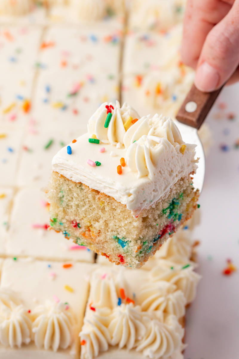a slice of frosted funfetti sheet cake on a cake server with the remaining cake in the background