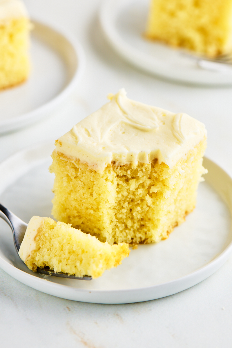 a slice of frosted vanilla sheet cake on a white plate with a bite on a fork