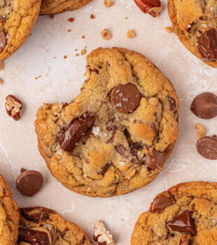 overhead close-up of a bakery-style chocolate chip cookie filled with two kinds of chocolate, toasted nuts, and toffee bits, plus a sprinkle of flaky salt on top
