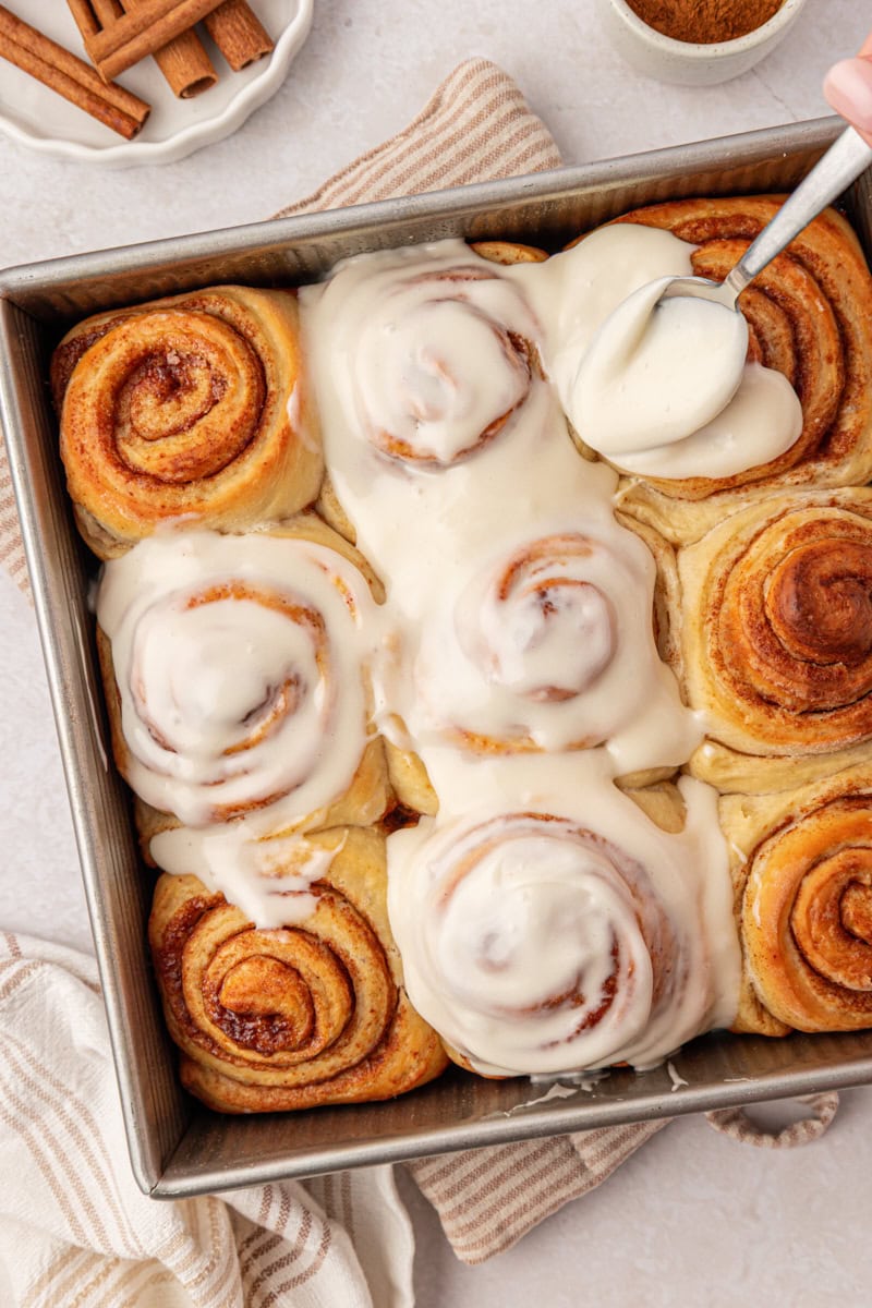 cream cheese frosting being spread over warm cinnamon rolls in a square baking pan