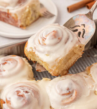 a cinnamon roll on a cake server being lifted away from the remaining rolls in a square baking pan