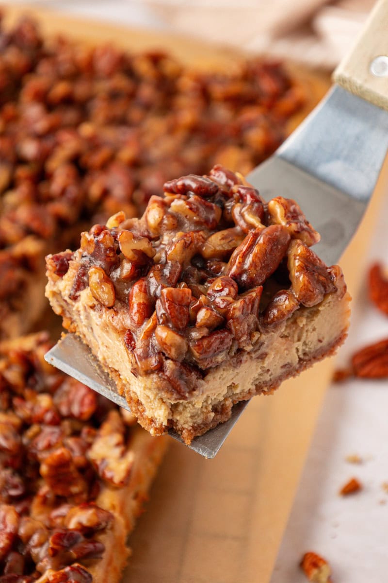 a pecan pie cheesecake on a serving spatula lifting away from the remaining bars