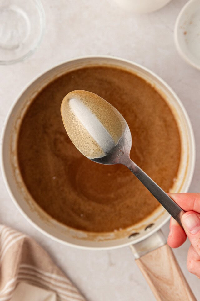 thick brown sugar caramel coating a spoon above a saucepan while making pecan pie cheesecake bar topping