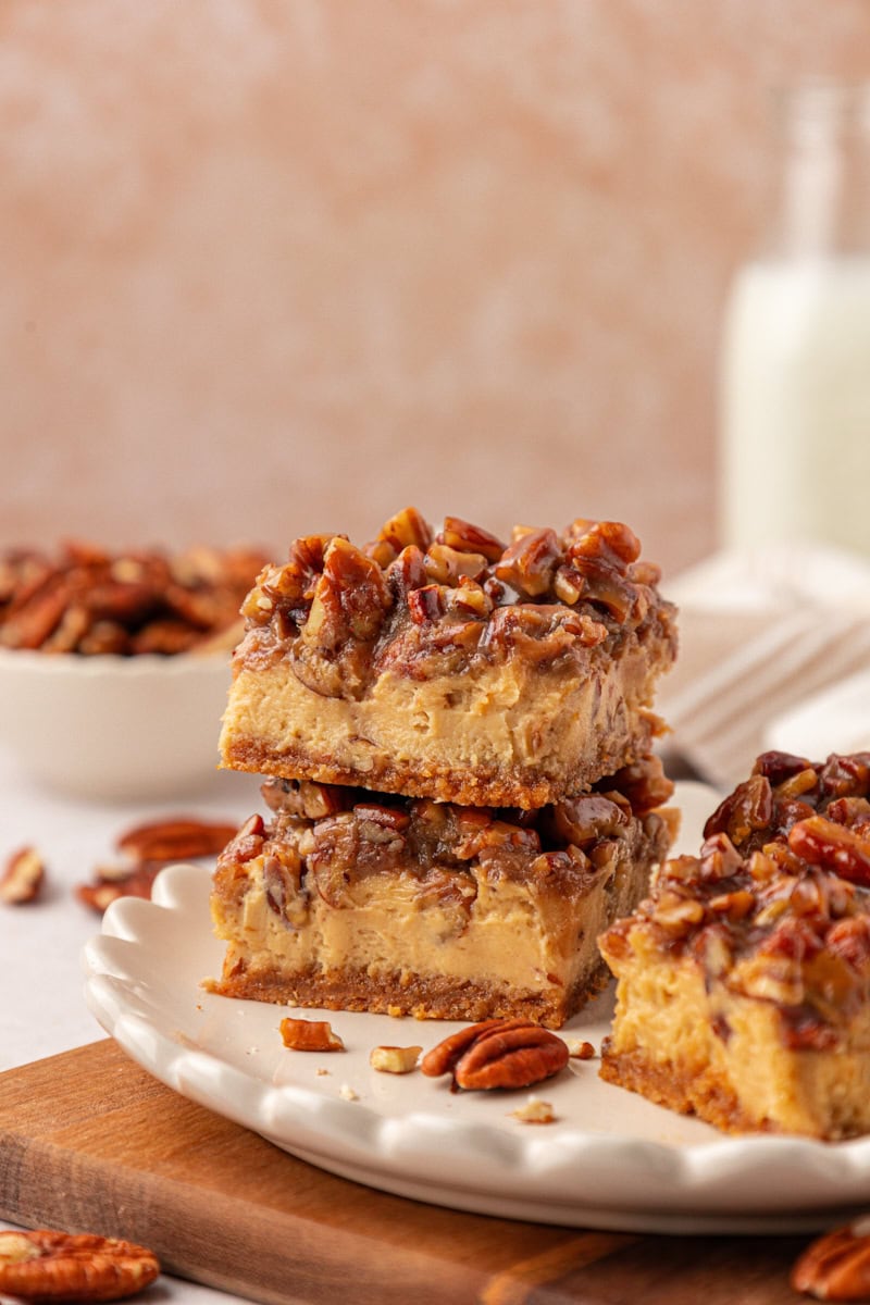 two stacked pecan pie cheesecake bars with layers of graham cracker crust, brown sugar cheesecake, and gooey pecan pie topping